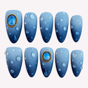 Stormlight blue ombre nails with glossy raised droplets and gold-tone frames around blue jewel-like accents.