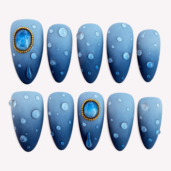 Stormlight blue ombre nails with glossy raised droplets and gold-tone frames around blue jewel-like accents.